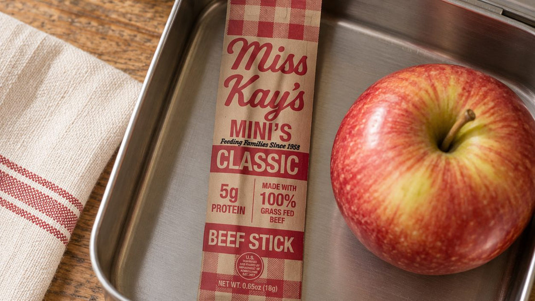 A Miss Kay's Mini's beef stick and a red apple in an open steel lunch box on a wooden table