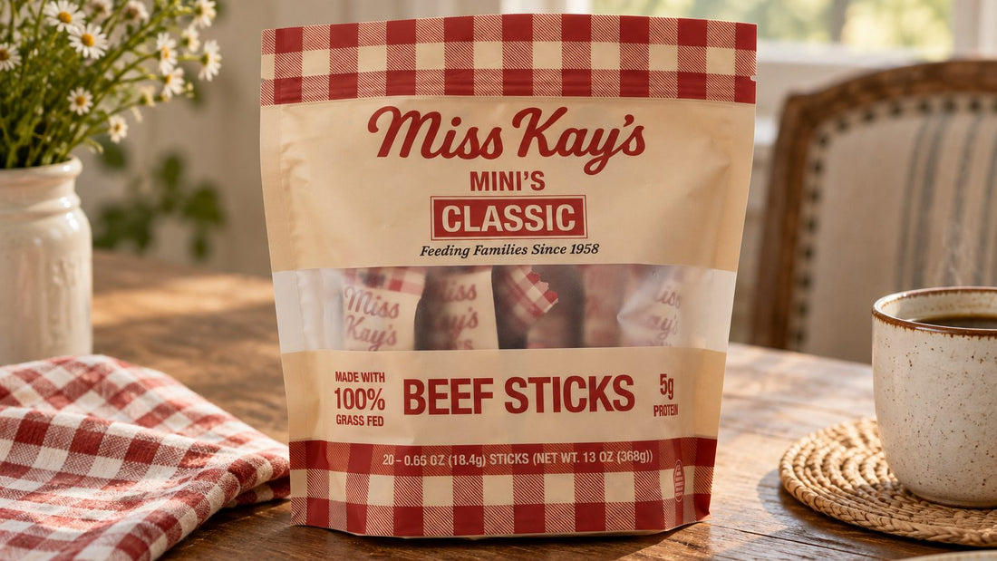 A bag of Miss Kay's Mini's Classic beef sticks on a wooden kitchen table beside a coffee cup and a gingham napkin