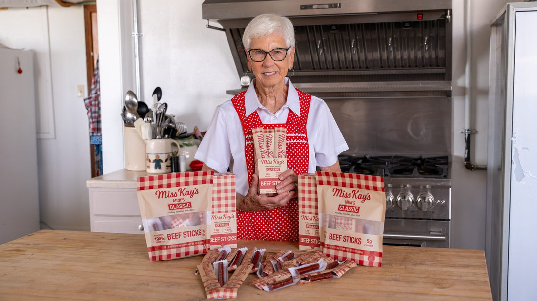 Miss Kay in her kitchen holding a box of Classic beef sticks, with bags, boxes and loose sticks on the counter