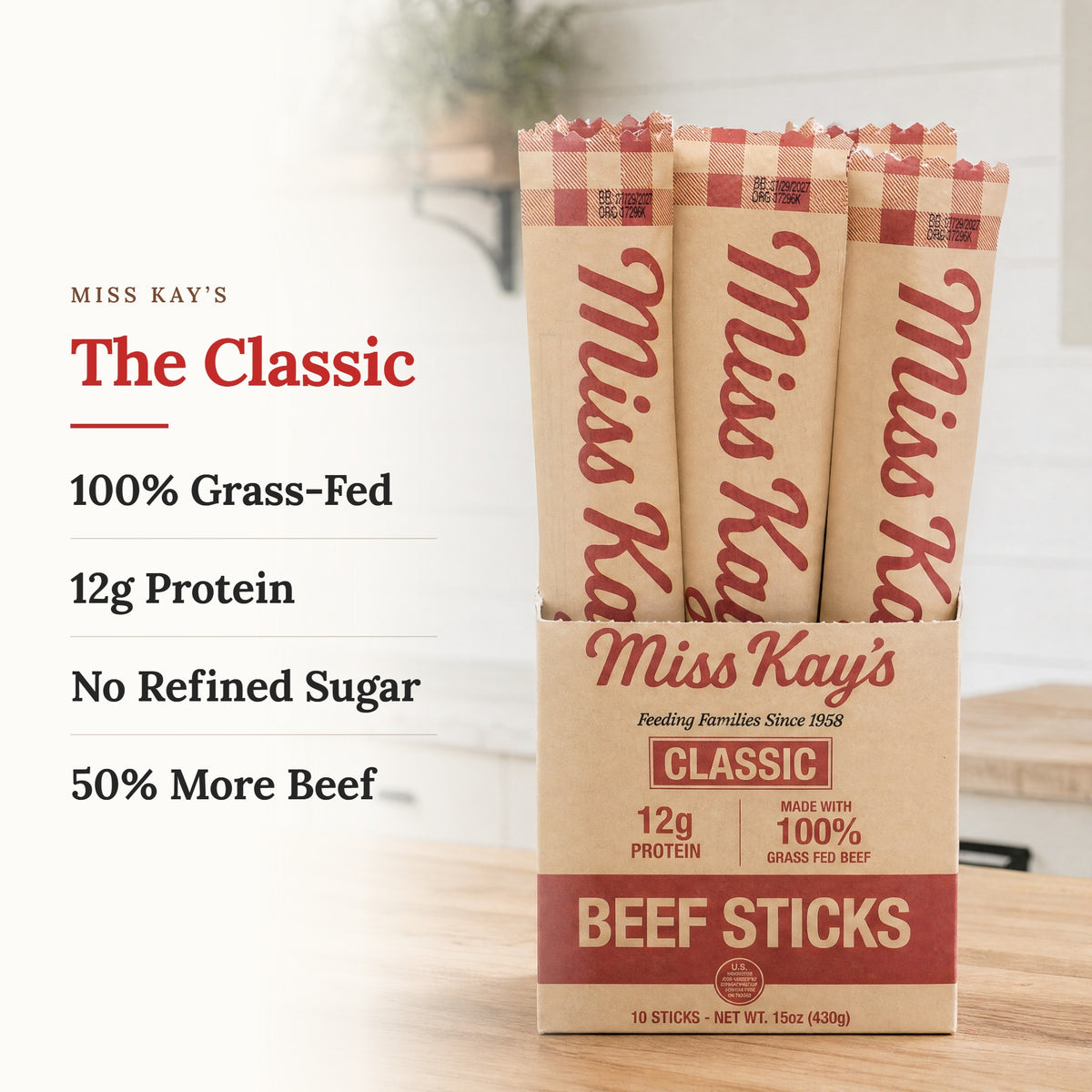 Miss Kay's Classic Beef Sticks displayed in a cardboard tray with three individual sticks standing behind it. The packaging is kraft paper with red text and stripes, set on a wooden surface with a blurred kitchen background.