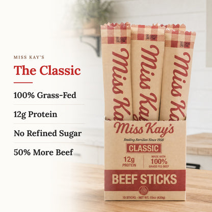 Miss Kay's Classic Beef Sticks displayed in a cardboard tray with three individual sticks standing behind it. The packaging is kraft paper with red text and stripes, set on a wooden surface with a blurred kitchen background.