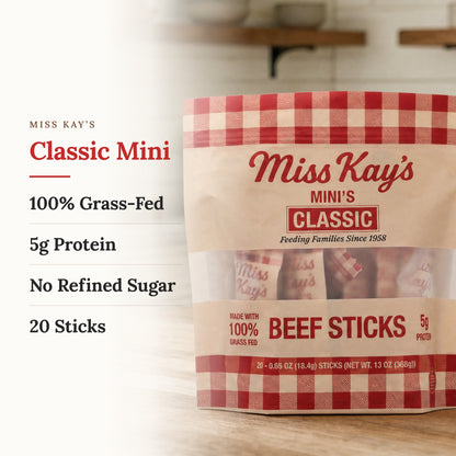 Miss Kay's Classic Mini Beef Sticks in a 13 oz bag, shown on a light wood surface with text callouts on the left detailing '100% Grass-Fed', '5g Protein', 'No Refined Sugar', and '20 Sticks'.