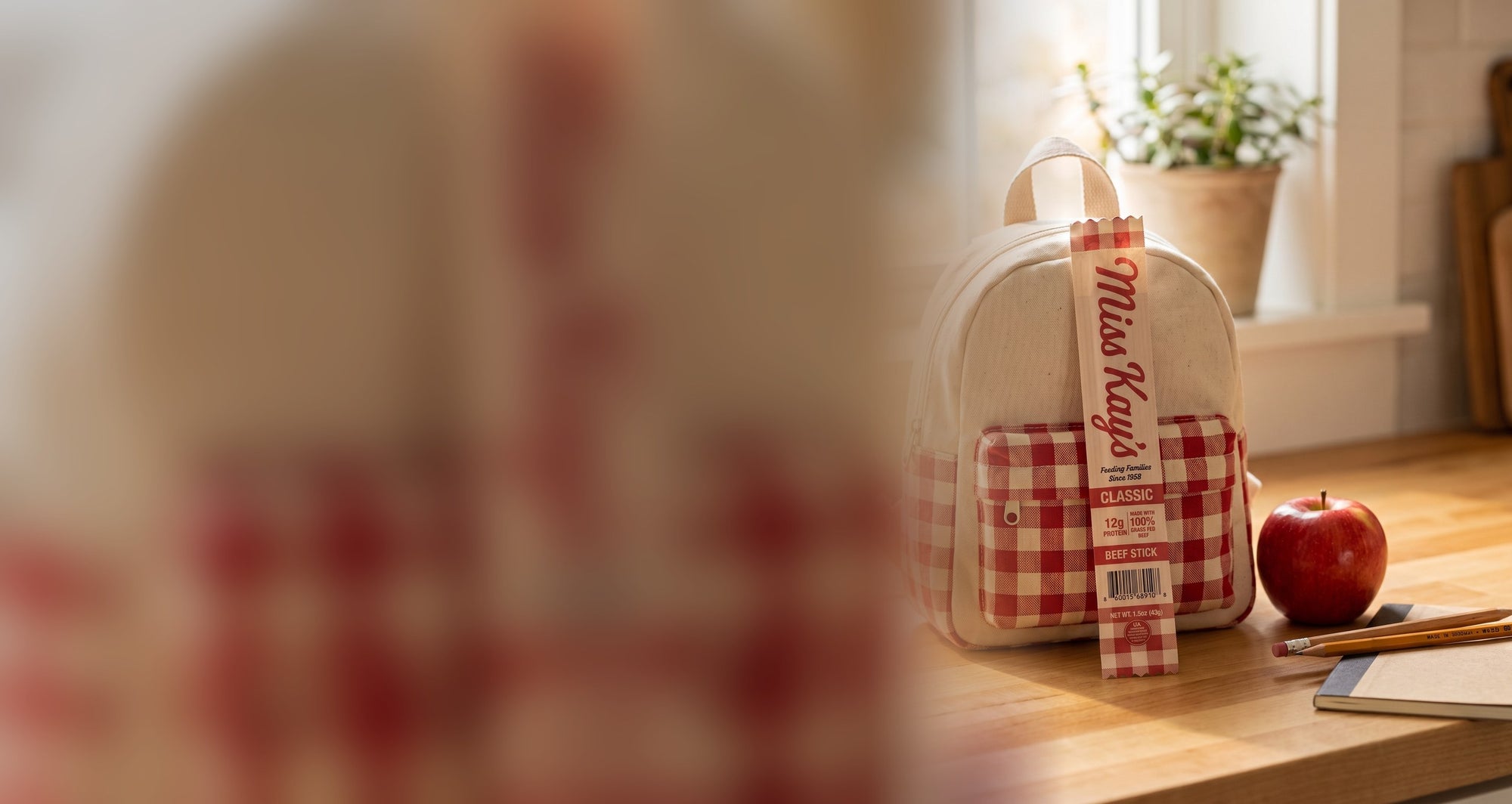 A Miss Kay's Classic beef stick tucked into a child's canvas backpack beside an apple and pencils on a sunlit kitchen table